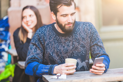 Man sitting at a cafe in Stockholm. He is typing on his smart phone and holding his coffee. Behind him there is a woman sitting at the table and looking away. Hipster and vintage look.